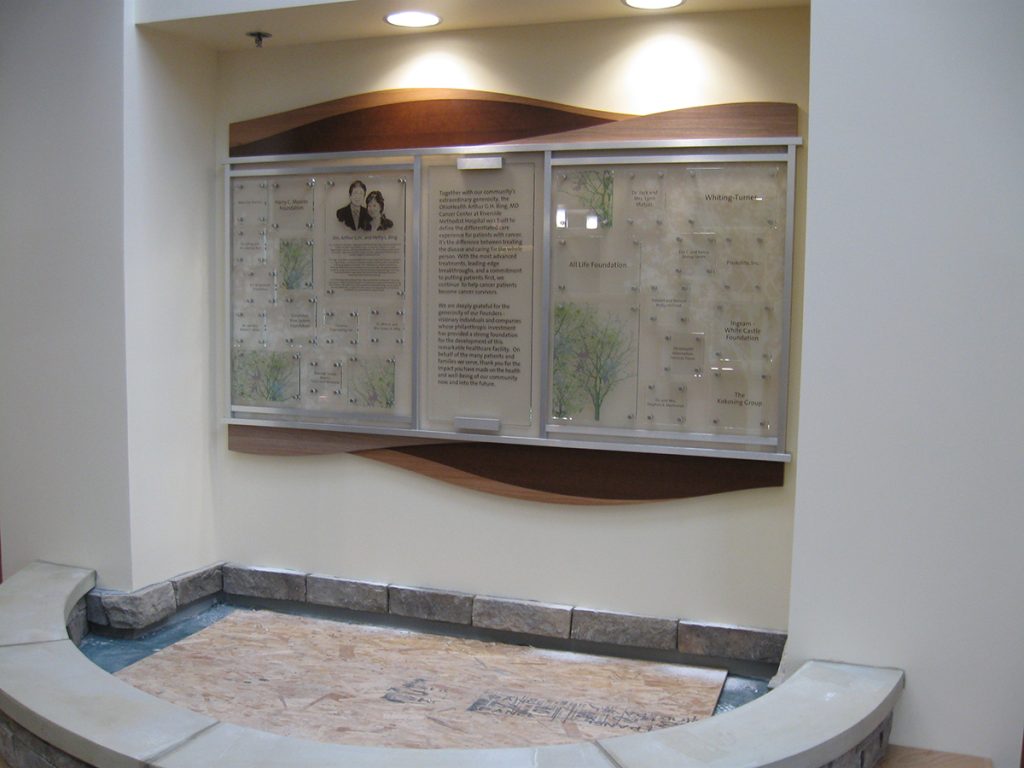 Close-up of the cancer center donor recognition display featuring flowing wood panels, glass donor nameplates mounted with satin silver stand-offs, and illustrated tree placeholder panels for future recognition.