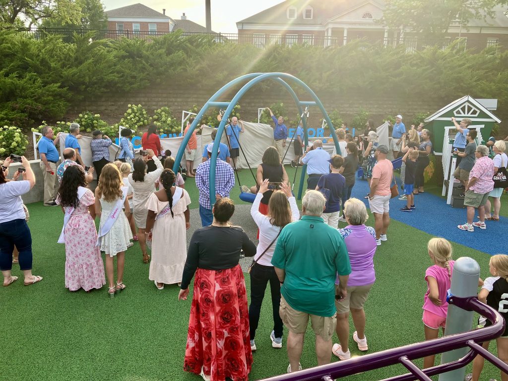 A large group of people—adults, children, and local dignitaries—have gathered around a covered donor display, which is being ceremoniously revealed. The event is celebratory, with many people taking photos or recording the moment on their phones.
