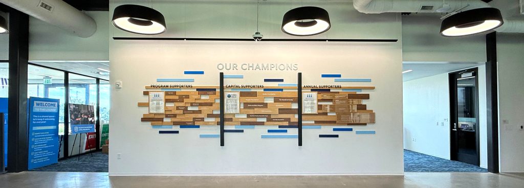 Freestanding white acrylic letters create a crisp title band above warm, wood toned laminates, with dimensional, vertical black accents setting rhythm and structure. High-gloss acrylic panels add depth and light - modern lines with a human feel - so the installation reads as both art and stewardship.