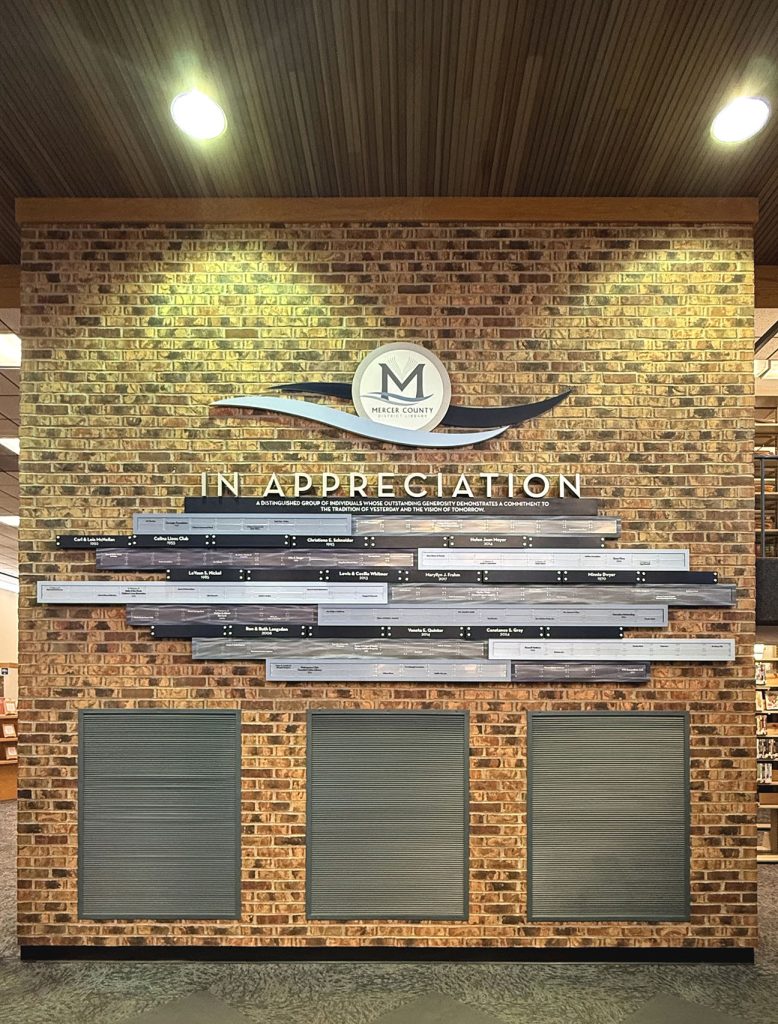 Unified donor recognition display at Mercer County District Library combining multiple plaques into one cohesive system on a brick wall.