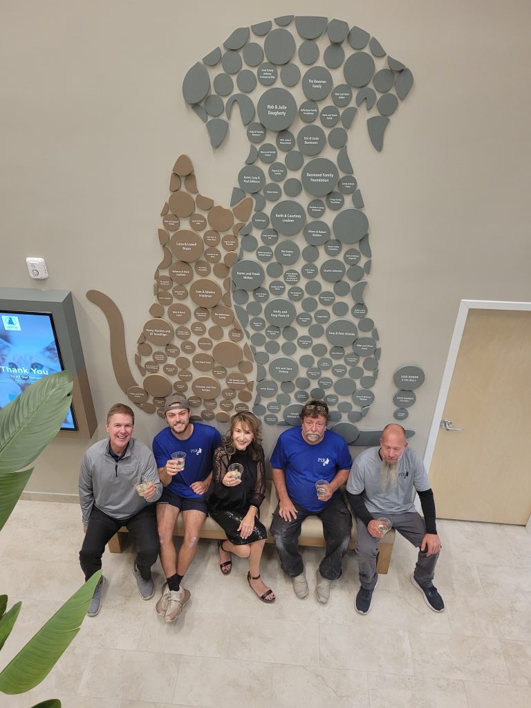 People seated in front of the completed donor recognition wall at ORCAT Grayvik Animal Center, showing the dog and cat silhouette design and real-world scale within the space.