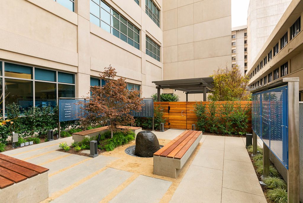 Outdoor memorial display integrated into a landscaped hospital garden with benches, stone feature, and blue-tinted memorial panels